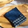 Mouchoir tissu double gaze artisanal plié avec soin, révélant le motif vichy bleu sur un plateau en bois ensoleillé.
