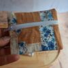 Gros plan sur un petit porte-monnaie artisanal bicolore associant un coton fleuri bleu et un tissu nid d'abeille ocre, tenu en main.