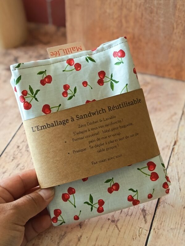 Emballage à sandwich réutilisable blanc à motif cerises rouges, entouré de son bandeau de présentation en papier kraft recyclé. Le bandeau affiche fièrement le nom du produit, la marque "Malili_fee" ainsi que les conseils d'utilisation pratiques.