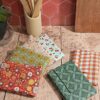 Vue d'ensemble de plusieurs emballages à sandwich réutilisables pliés en coton enduit, posés sur une table en bois dans une ambiance cuisine. On distingue différents motifs : floral rouge, géométrique vert, vichy orange et motif cerises. En arrière-plan, un pot avec des ustensiles en bois et un mur carrelé rouge.