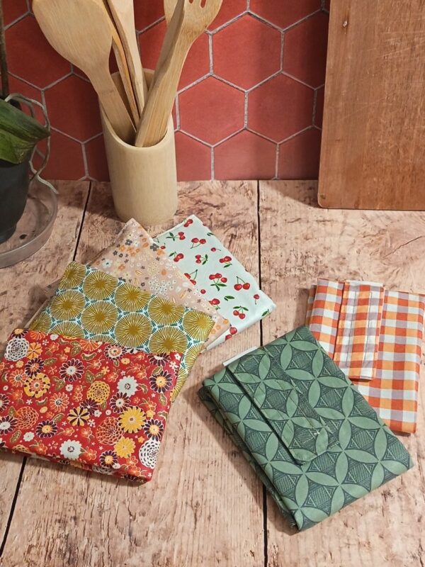 Vue d'ensemble de plusieurs emballages à sandwich réutilisables pliés en coton enduit, posés sur une table en bois dans une ambiance cuisine. On distingue différents motifs : floral rouge, géométrique vert, vichy orange et motif cerises. En arrière-plan, un pot avec des ustensiles en bois et un mur carrelé rouge.