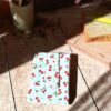 Emballage à sandwich réutilisable plié en coton enduit blanc avec un motif de petites cerises rouges. Il est posé sur une table en bois ensoleillée à côté d'une planche avec du pain de mie.