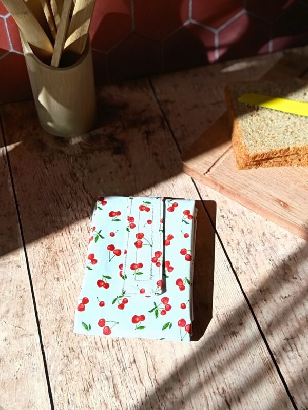 Emballage à sandwich réutilisable plié en coton enduit blanc avec un motif de petites cerises rouges. Il est posé sur une table en bois ensoleillée à côté d'une planche avec du pain de mie.