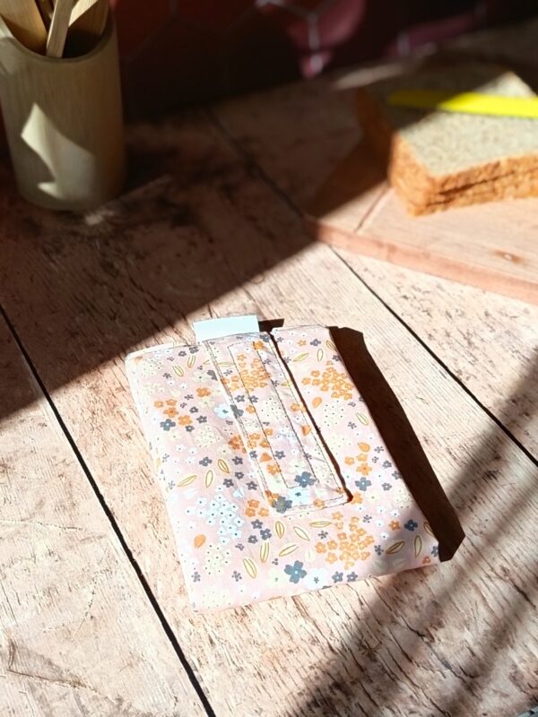 Zoom sur la variation de l'emballage à sandwich réutilisable en tissu floral rose poudré et saumon. Le produit est fermé par sa bande scratch et présenté dans un décor de cuisine lumineux.