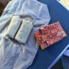 Vue du dessus d'un emballage à sandwich réutilisable entièrement déplié sur une table de camping bleue. Il est utilisé comme set de table et contient deux wraps bien garnis prêts à être dégustés. Sa doublure intérieure en film EVA certifié contact alimentaire est visible. L'emballage pliable en coton enduit rouge à motifs floraux est posé à côté.