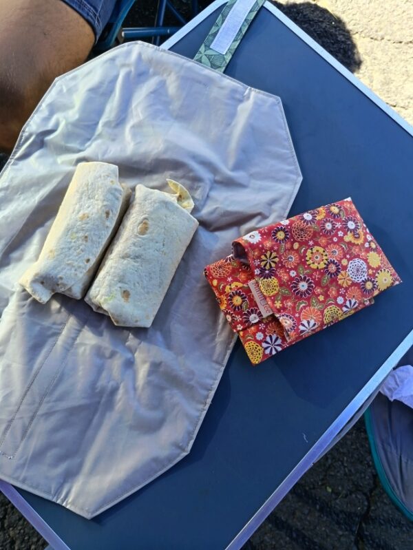 Vue du dessus d'un emballage à sandwich réutilisable entièrement déplié sur une table de camping bleue. Il est utilisé comme set de table et contient deux wraps bien garnis prêts à être dégustés. Sa doublure intérieure en film EVA certifié contact alimentaire est visible. L'emballage pliable en coton enduit rouge à motifs floraux est posé à côté.