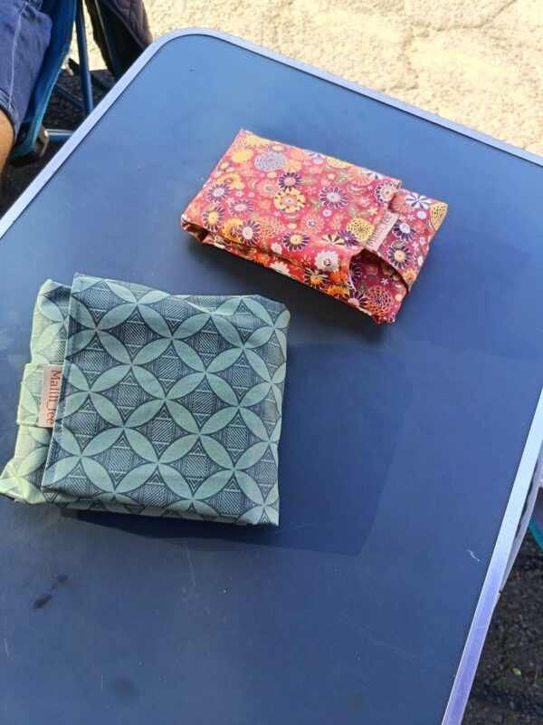 Gros plan sur deux emballages à sandwich réutilisables pliés, posés côte à côte sur une table de camping bleue. L'un est en coton enduit rouge à motif floral coloré, l'autre est vert avec un motif géométrique discret. On voit clairement les étiquettes en tissu "Malili_fee" cousues sur chaque emballage.