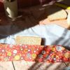 Démonstration du pliage d'un emballage à sandwich réutilisable à motif floral rouge. Le premier rabat en tissu enduit recouvre une partie de la tranche de pain de mie, laissant apparaître la doublure intérieure grise étanche et la planche en bois en arrière-plan.