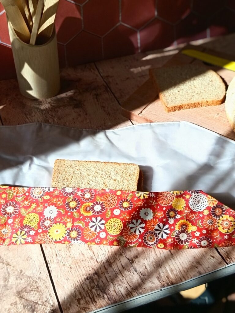 Démonstration du pliage d'un emballage à sandwich réutilisable à motif floral rouge. Le premier rabat en tissu enduit recouvre une partie de la tranche de pain de mie, laissant apparaître la doublure intérieure grise étanche et la planche en bois en arrière-plan.