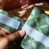 Une main présente le système de fermeture d'un emballage à sandwich lavable en tissu vert. On observe les larges bandes de scratch blanc auto-agrippant, idéales pour ajuster la taille de la pochette selon le format du repas.