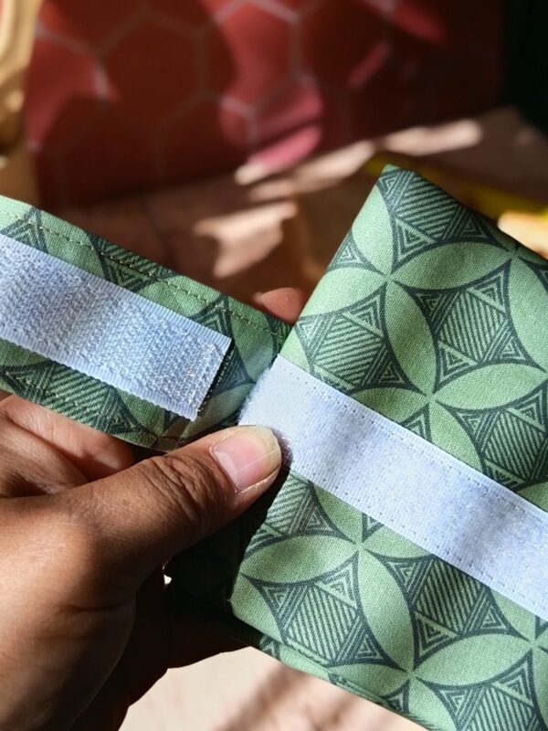 Une main présente le système de fermeture d'un emballage à sandwich lavable en tissu vert. On observe les larges bandes de scratch blanc auto-agrippant, idéales pour ajuster la taille de la pochette selon le format du repas.