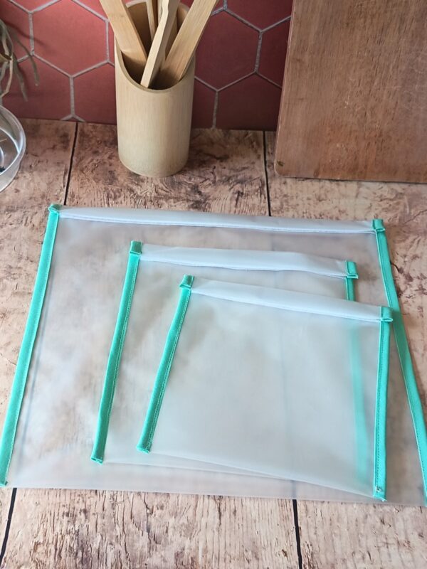 Présentation à plat d'un lot de trois sacs de congélation réutilisables de tailles différentes. Le film EVA translucide laisse deviner le plan de travail en bois tandis qu'un biais de couleur vert menthe borde soigneusement chaque modèle.