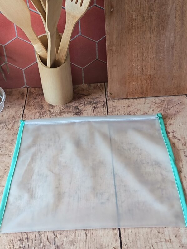 Présentation du grand sac de congélation réutilisable de taille L ($33 \times 30\text{ cm}$) bordé d'un biais vert menthe, posé à plat dans une cuisine devant des ustensiles en bois.
