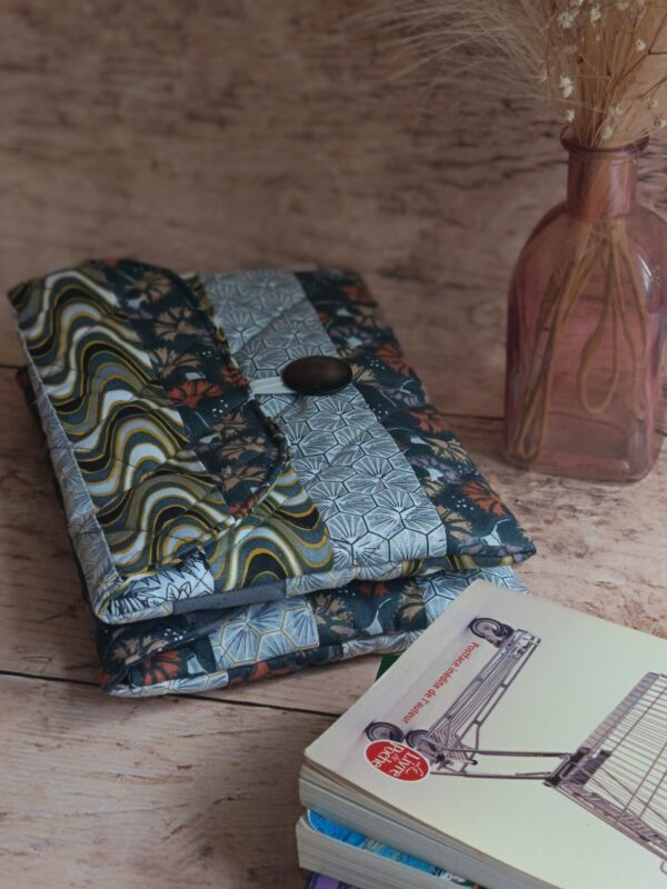 Housse de livre de poche en tissu matelassé gris aux motifs japonais et vagues, posée près d'un livre et d'un soliflore.