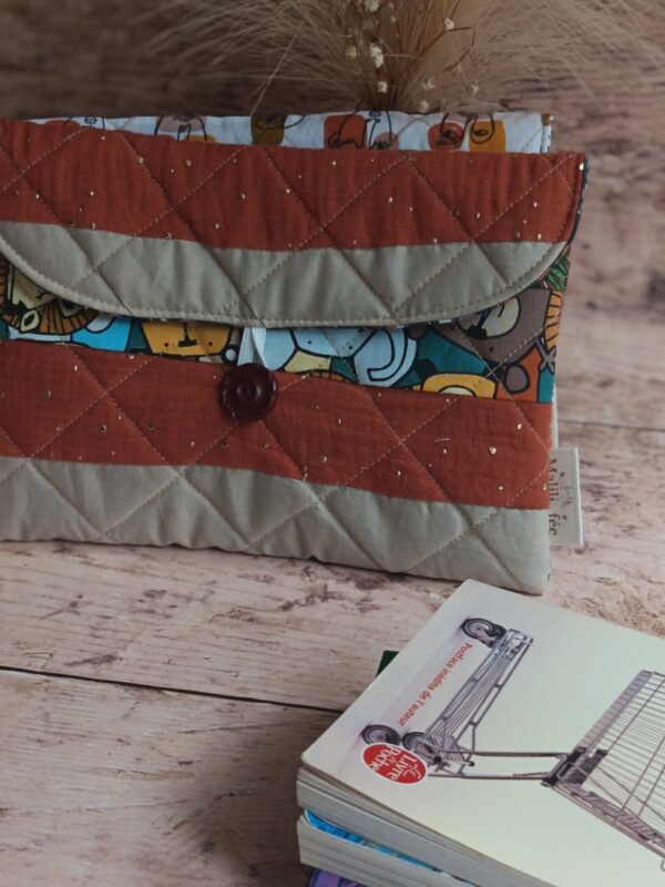 Pochette à livre de poche écoresponsable en tissu matelassé terracotta et beige, posée près de livres sur une table en bois.