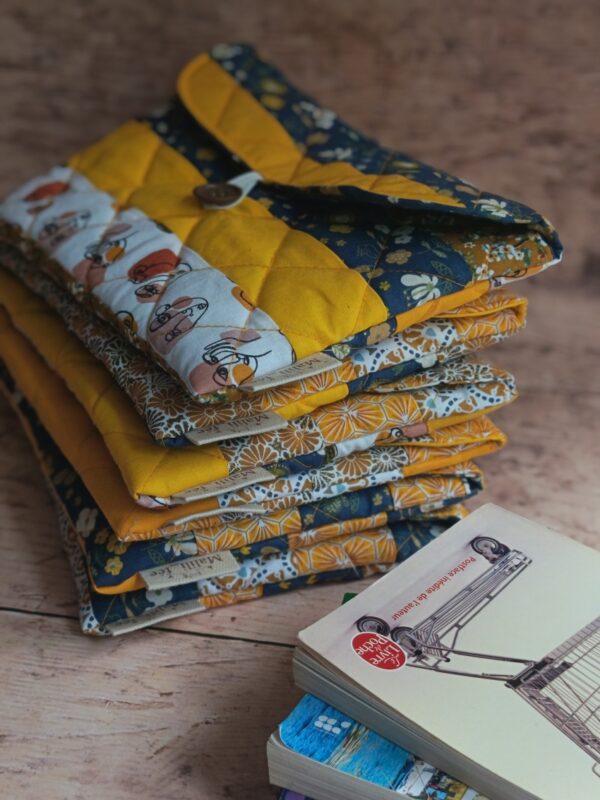 Pochette à livre de poche en tissu matelassée jaune moutarde et bleu nuit, posée sur une pile avec des livres.