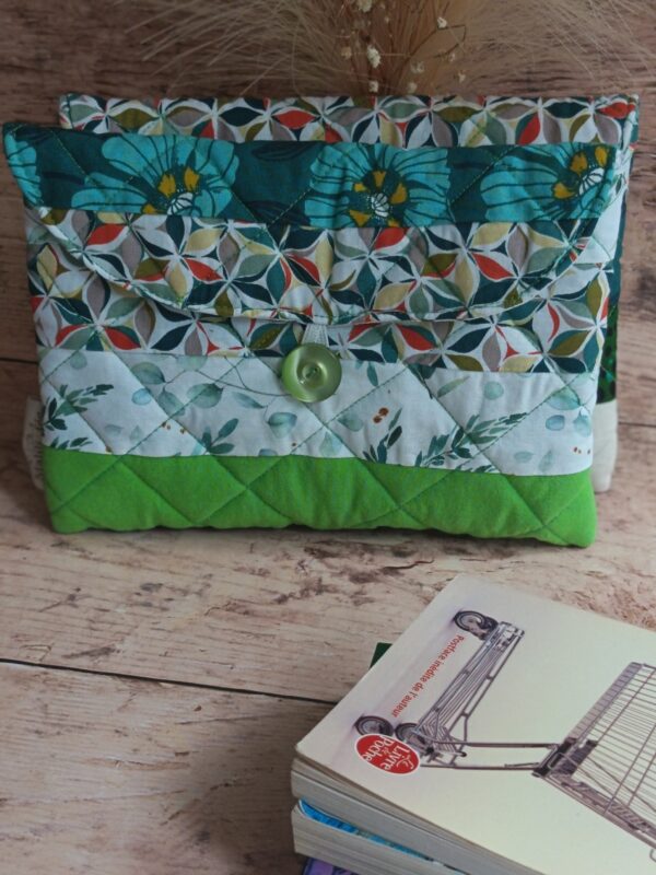 Pochette à livre de poche faite main en tissu matelassé vert et motifs feuillage avec bouton de fermeture et pile de livres.