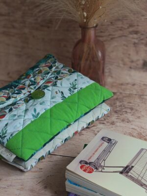 Pochette à livre de poche faite main matelassée aux motifs végétaux vert pomme et vert canard, posée près de livres.