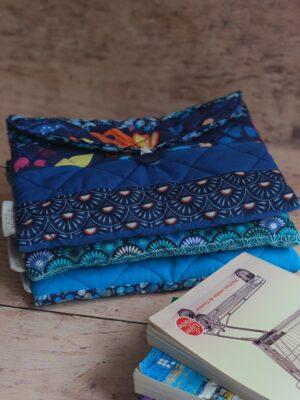 Pile de trois pochettes à livre de poche en tissu matelassé aux nuances de bleu, posée à côté d'un livre de poche.