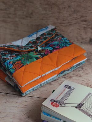 Pochette à livre de poche originale en tissu matelassé orange et motifs tropicaux, posée à côté d'un livre.