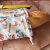Baguette fraîche dépassant d'un sac à baguette nomade en coton motif boulangerie rétro fermé par un cordon blanc.