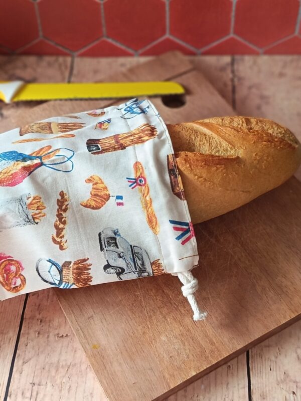 Baguette fraîche dépassant d'un sac à baguette nomade en coton motif boulangerie rétro fermé par un cordon blanc.